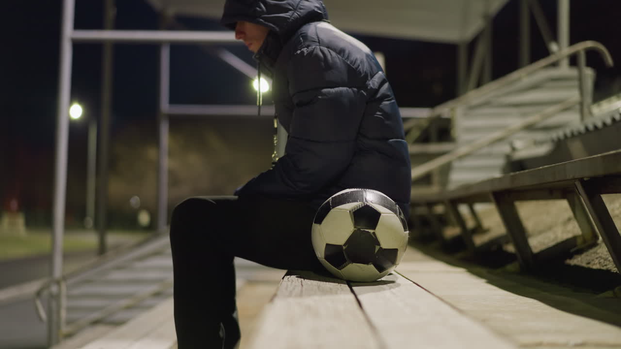 Close-up side view of a man in a black outfit and jacket sitting alone in a quiet, dimly lit stadium, he is seated with his hands between his legs, with a soccer ball next to him