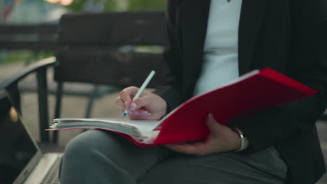 close up of woman in professional dress reviewing documents in red folder with pen in hand while seated outdoors beside laptop with blurred background featuring benches and greenery