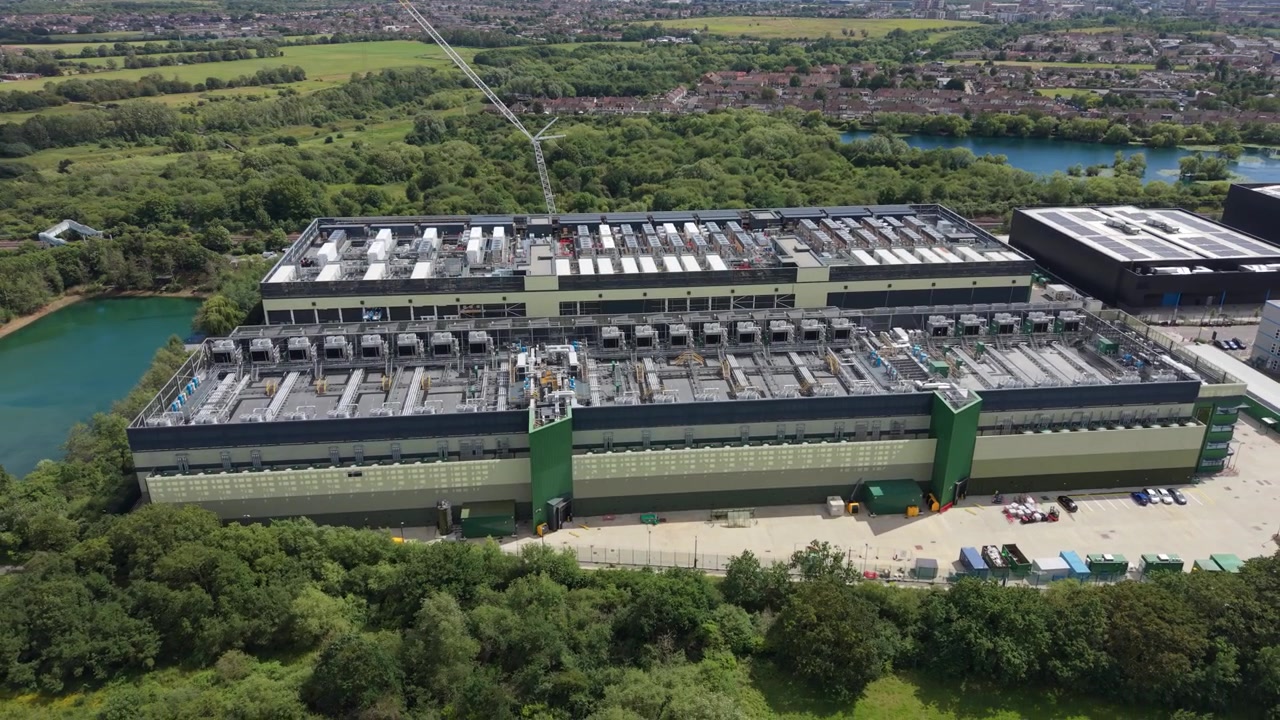 This shows an aerial perspective of a massive data center campus with multiple large buildings. It is situated within a lush green landscape, featuring trees, grass, and nearby water bodies.