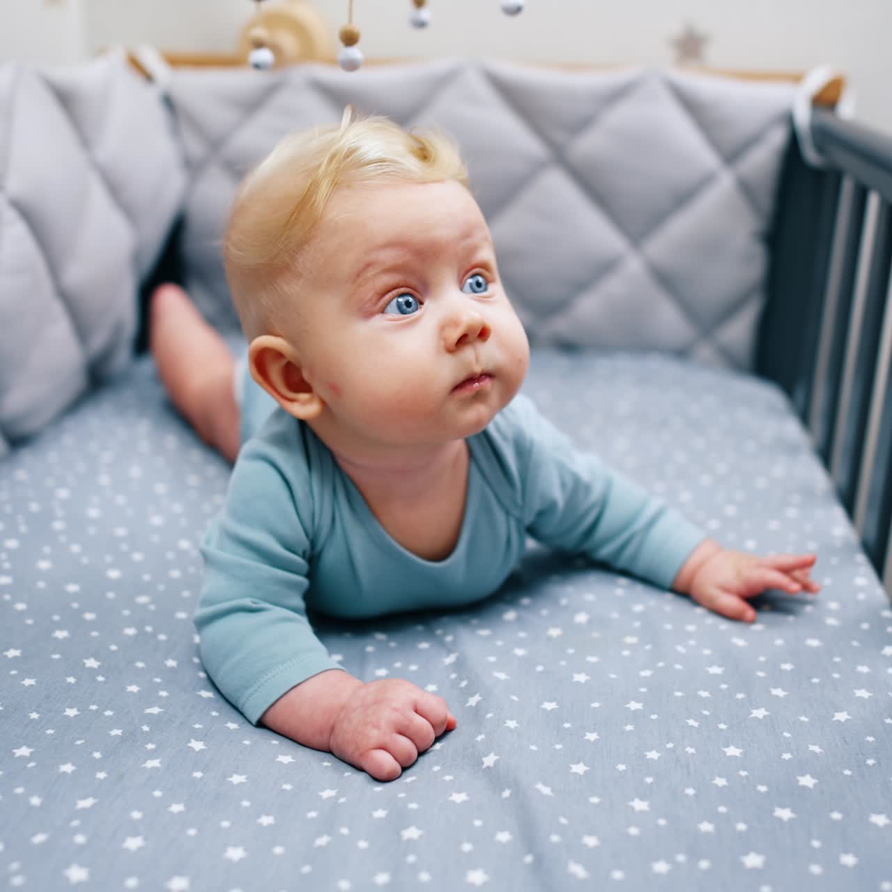 Liten babygutt med store blå øyne ligger på magen. Søt baby iført blå body i sengen.