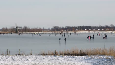 people ice skating on a frozen lake past a windmill on a sunny winter day in the netherlands during the pandemic