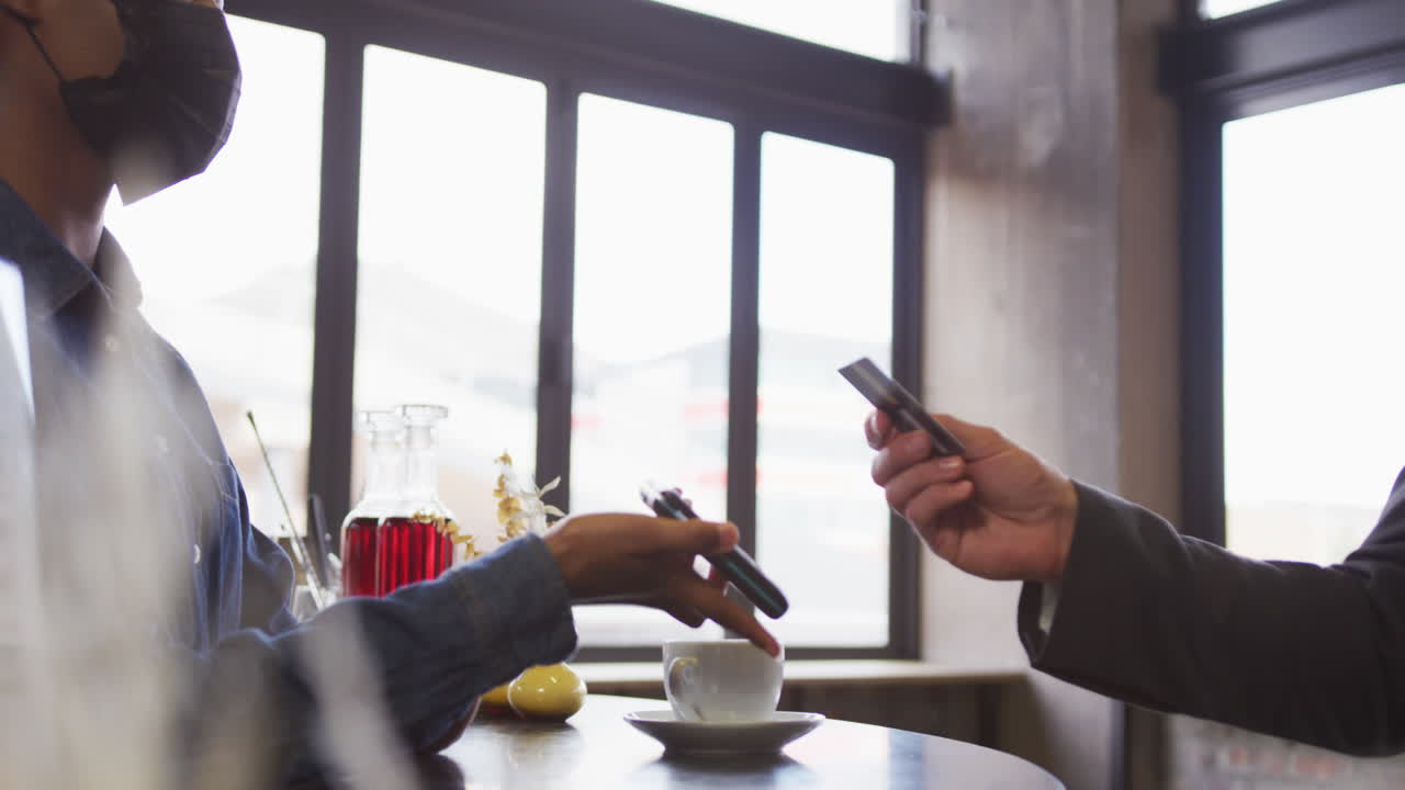 African american woman in face mask taking contactless card payment from customer at cafe bar