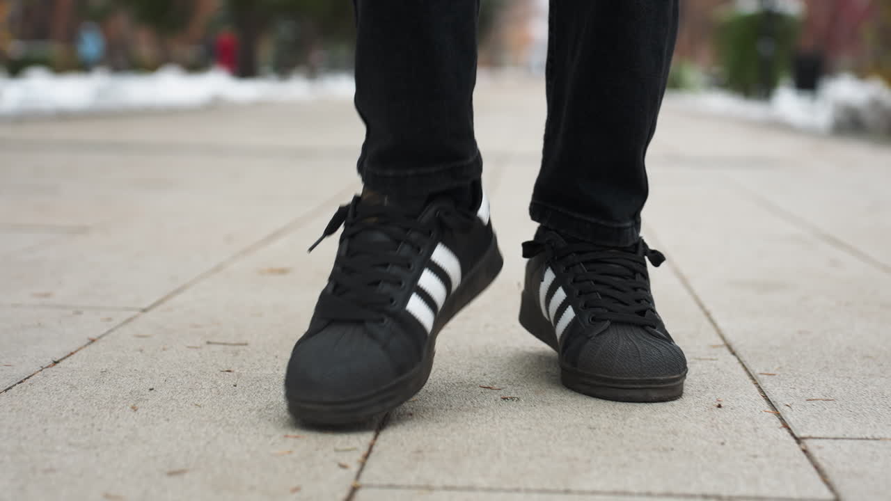 검은색 바지를 입고 색 줄무를 가진 검은색 운동화를 입은 사람이 포장된 교차된 길을 고, 배경은 눈, 초록색 및 도시 설정의 암시로 흐릿합니다.