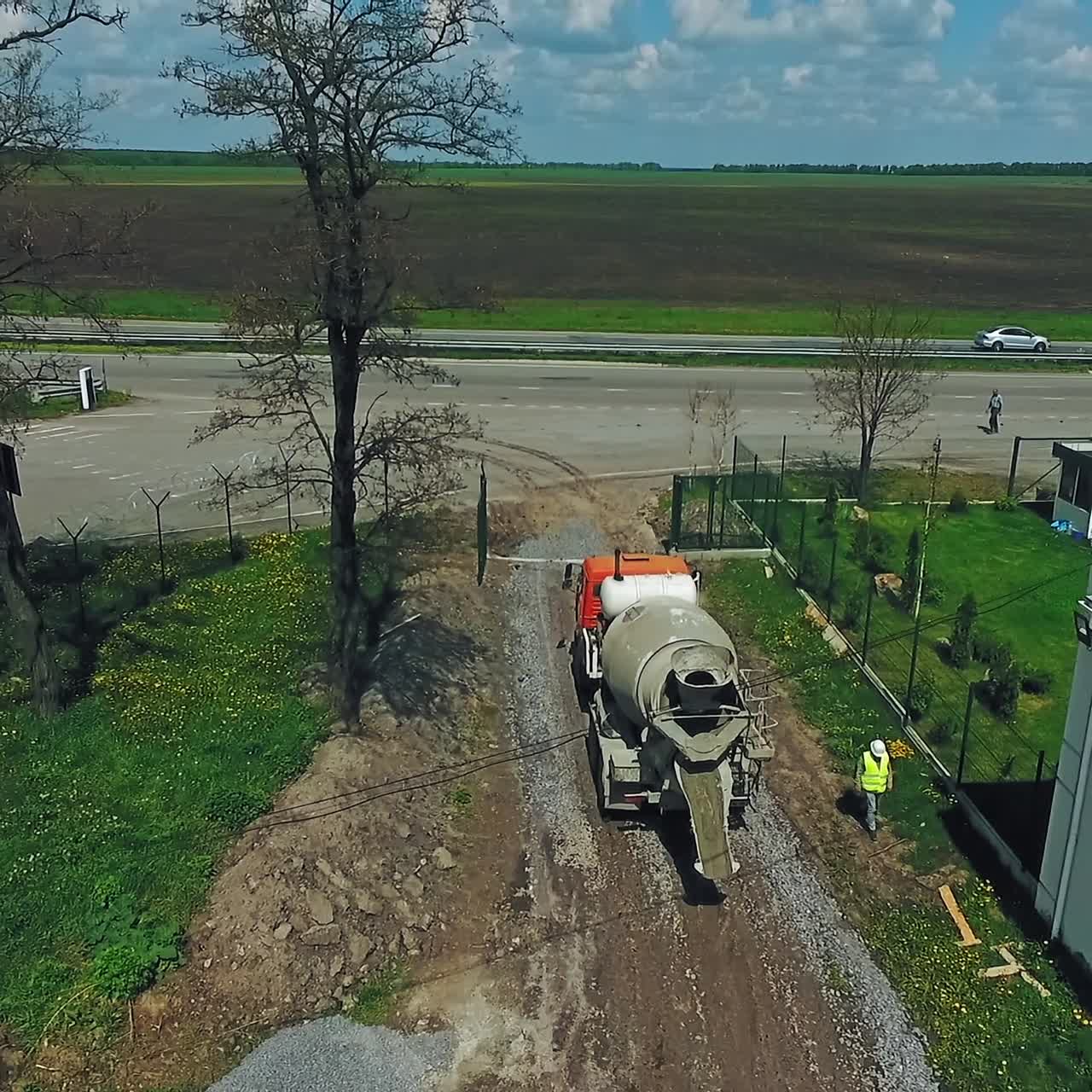 Concrete mixing machine moving by the road covered with gravel. Worker in vest and helmet walking next to the car. Highway and agricultural field at backdrop