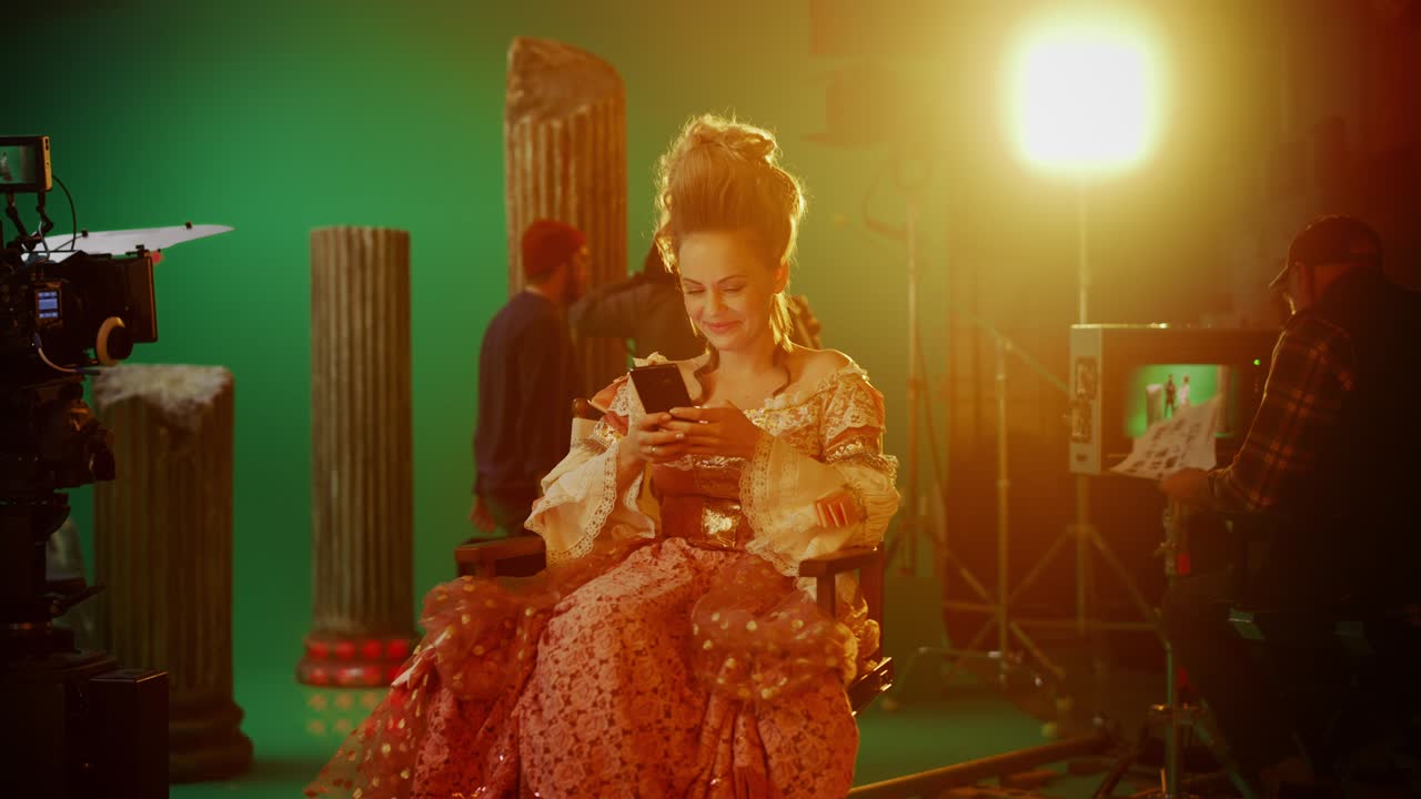 en el set de la película de drama de época: hermosa actriz sonriente con un vestido renacentista, sentada en una silla usando un teléfono inteligente con pantalla verde en el fondo. estudio de rodaje de éxito de taquilla histórico
