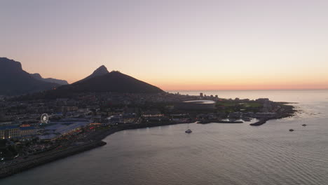 Aerial-panoramic-footage-of-sea-coast-in-city-at-dusk.-Buildings-on-waterfront-and-hills-against-colourful-sky.-Cape-Town,-South-Africa