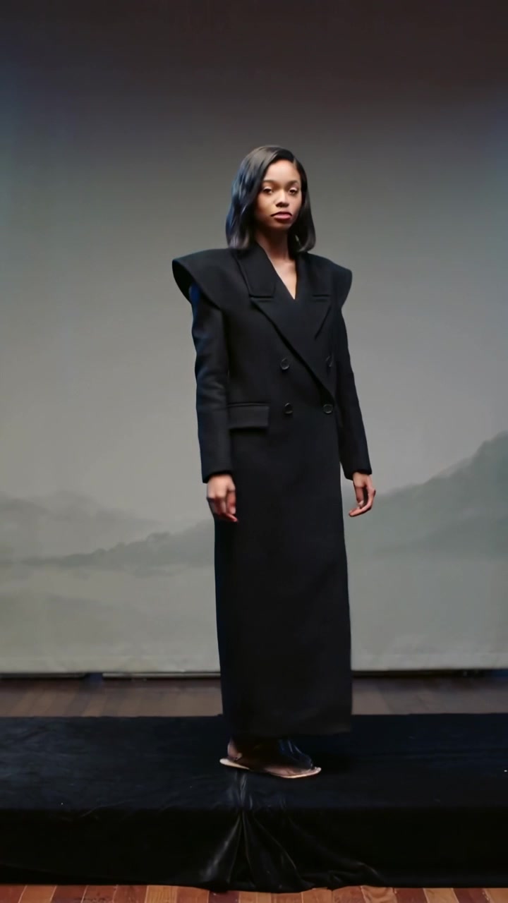 Lodret video: Model drejer i takt og præsenterer lang frakke med markerede skuldre på lav catwalk.