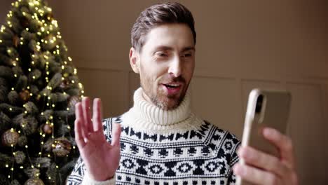 close up portrait of happy male in good mood videochatting on cellphone near glowing decorated xmas tree