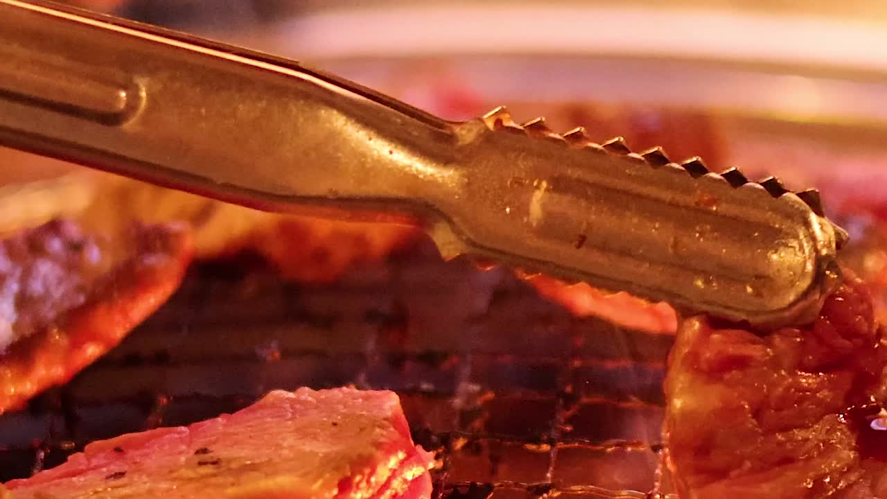 Fettine sottili di carne rossa sfrigolano su una griglia a carbone mentre delle pinze metalliche le girano.