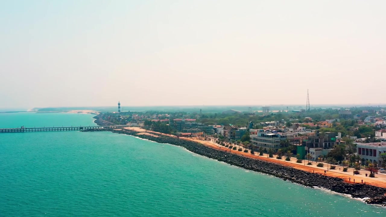La Goubert Beach Road di Puducherry si estende lungo la costa del Golfo del Bengala con acqua turchese e sviluppo urbano. Una carrellata fluida rivela il lungomare e il paesaggio urbano sotto una luce solare soffusa