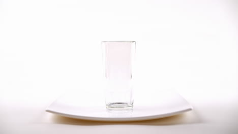 mano sumergiendo una bolsa de té en un vaso claro de agua en un plato blanco, fondo blanco