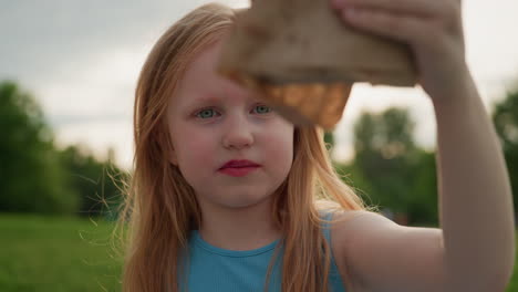 portrait creative girl waving pastry wrapped in brown paper in sunny recreational park smiling gently while enjoying playful snack time with nature background and soft evening light