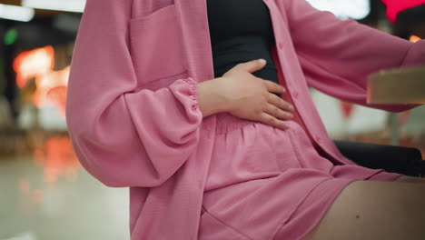 woman wearing a pink dress gently rubbing her stomach with her right hand, resting the other hand on the table after a meal, with blurred lights and people walking in the background