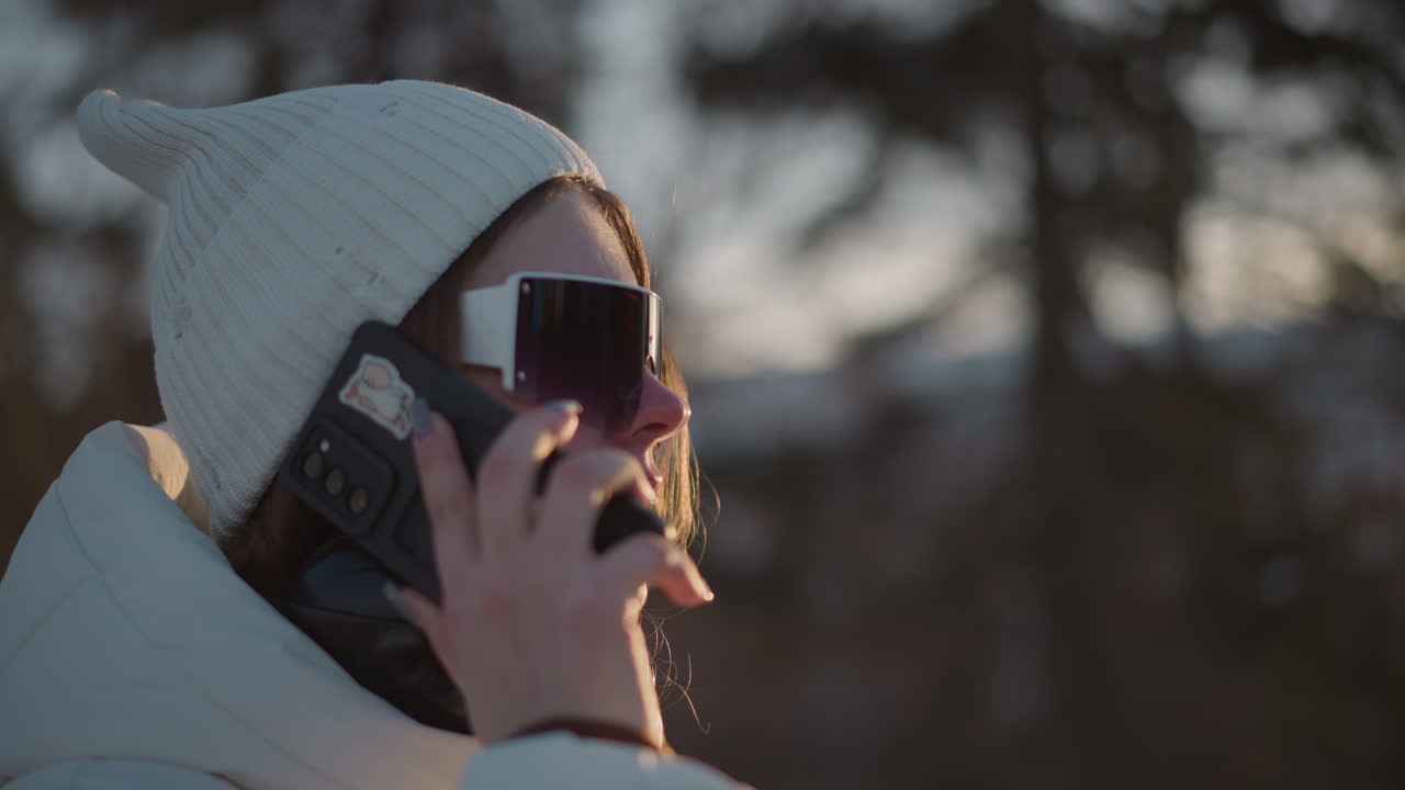 Side view of young girl holding phone to ear talking and smiling while shaking body under warm sunset light wearing white beanie and puffer coat against snowy forest background