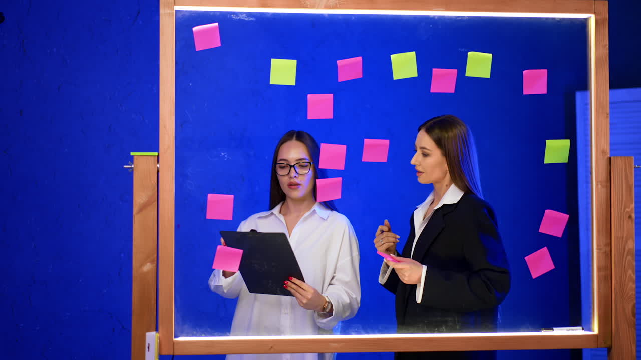 Women teamwork with sticky notes. Two women discuss ideas in front of a glass board covered with colorful sticky notes during a brainstorming session