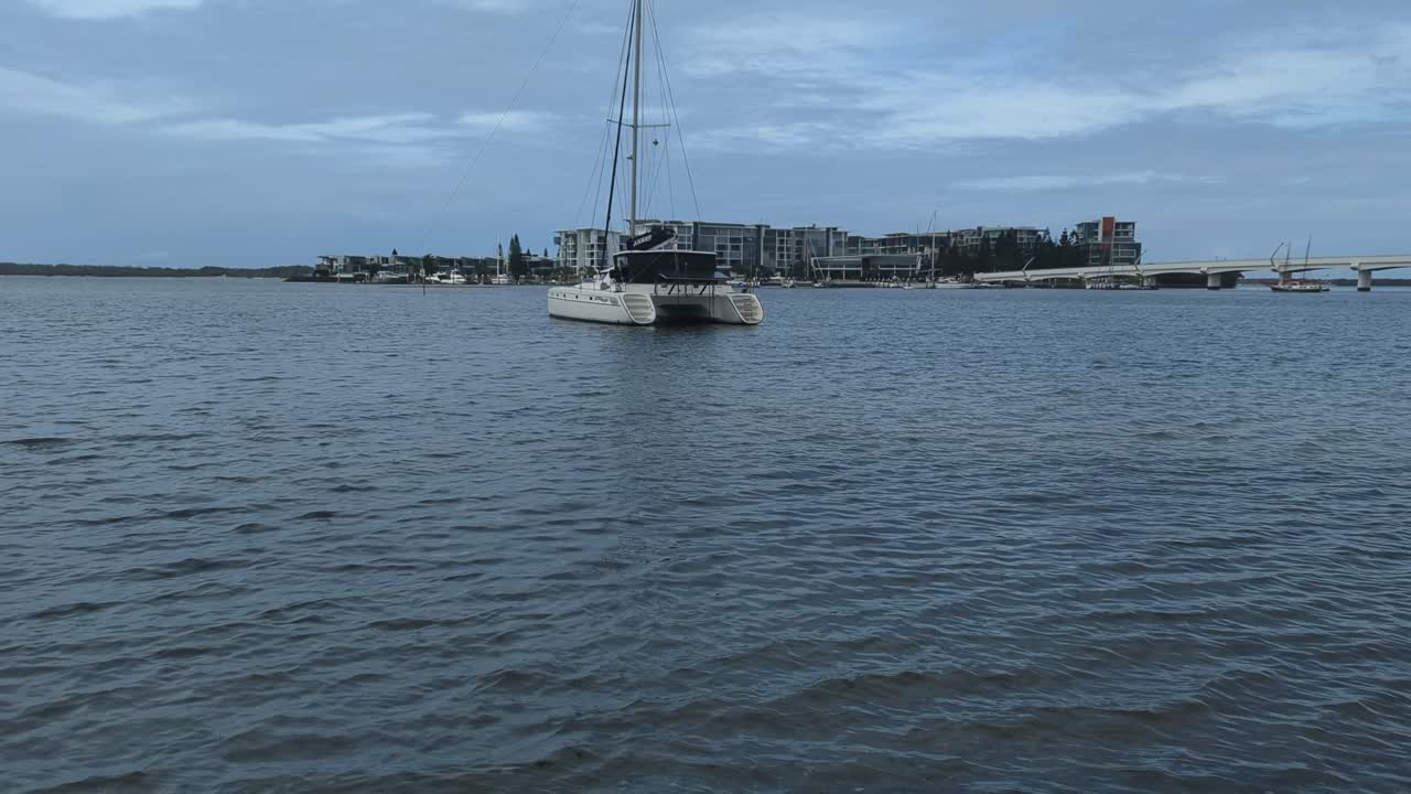 Pan up, A Catamaran anchored in the shallow waters of a beach with an island resort and bridge in the background.