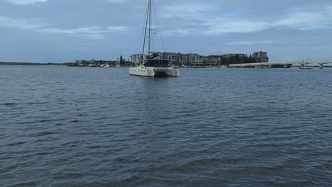 pan up, a catamaran anchored in the shallow waters of a beach with an island resort and bridge in the background.