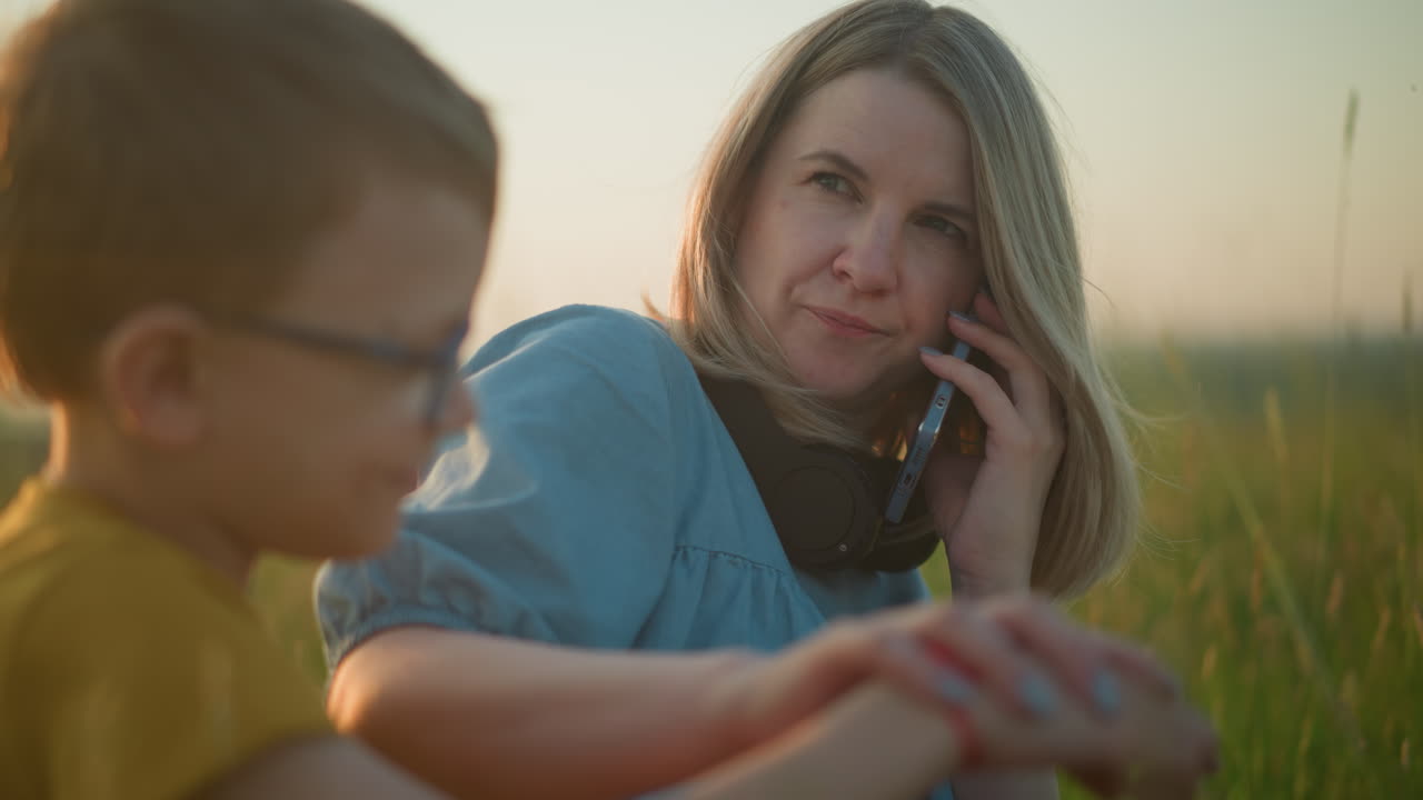 파란 옷을 입은 여자가 잔디에 앉아서 귀에 전화를 들고, 안경을 쓴 아이를 향해 미소 짓고, 에 흐릿하게 앉아, 야외 환경에서 따뜻하고 부드러운 순간을 만니다.