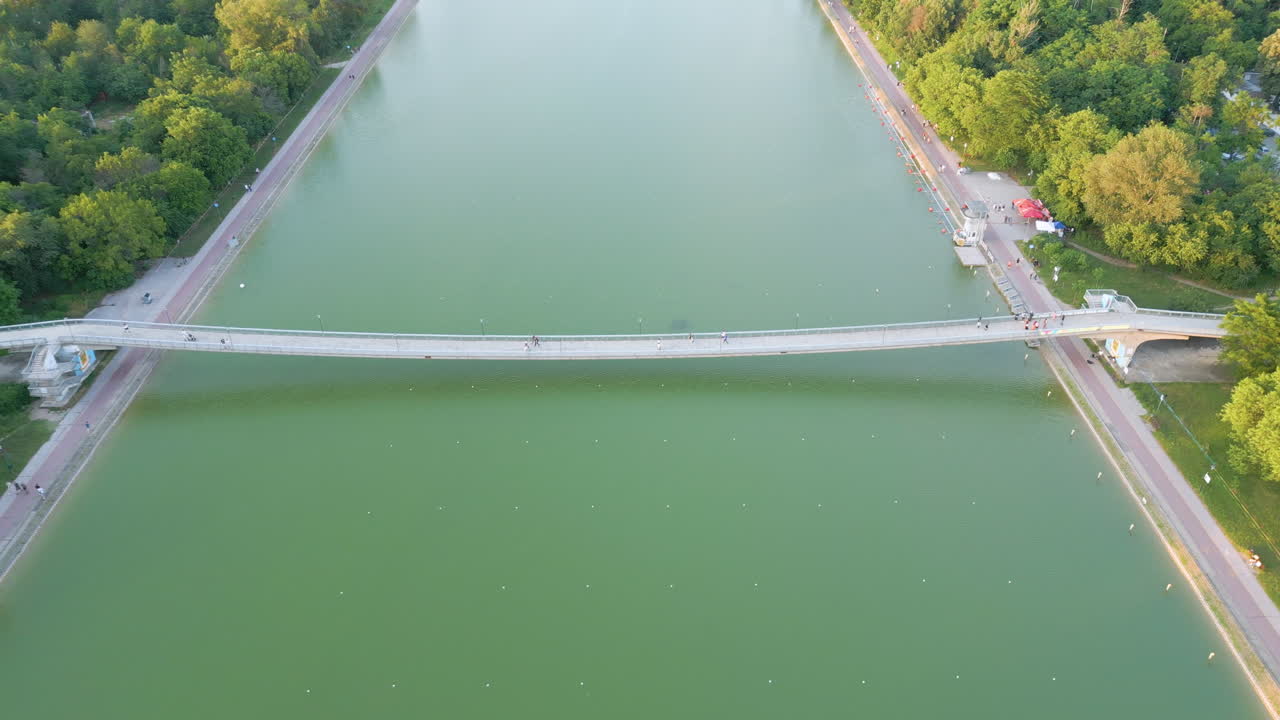 Top-down aerial pullback reveals the pedestrian bridge over Plovdiv’s rowing canal at sunset—calm green water with training buoys, soft clouds, and the city skyline unfolding in the distance
