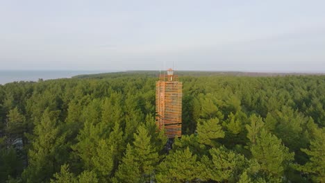 Aerial view of Bernati lighthouse surrounded by lush green pine tree forest with light snow, Nordic woodland, Baltic sea coast, sunny winter day, Latvia, wide ascending drone shot moving backward
