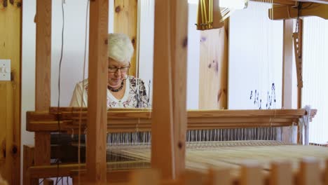 Front-view-of-old-caucasian-senior-woman-weaving-cloth-on-handloom-machine-in-a-workshop-4k