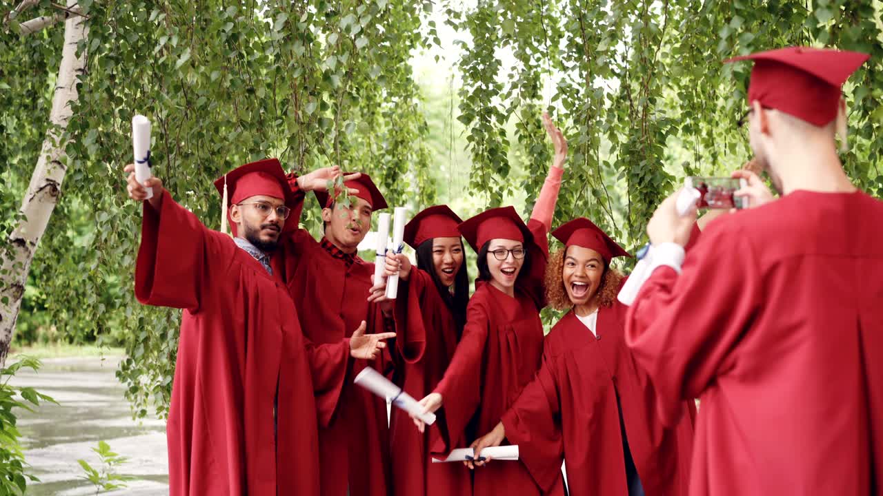 Graduating student is recording video of his friends in gowns holding diplomas, waving hands and posing looking at smartphone camera. People and technology concept.