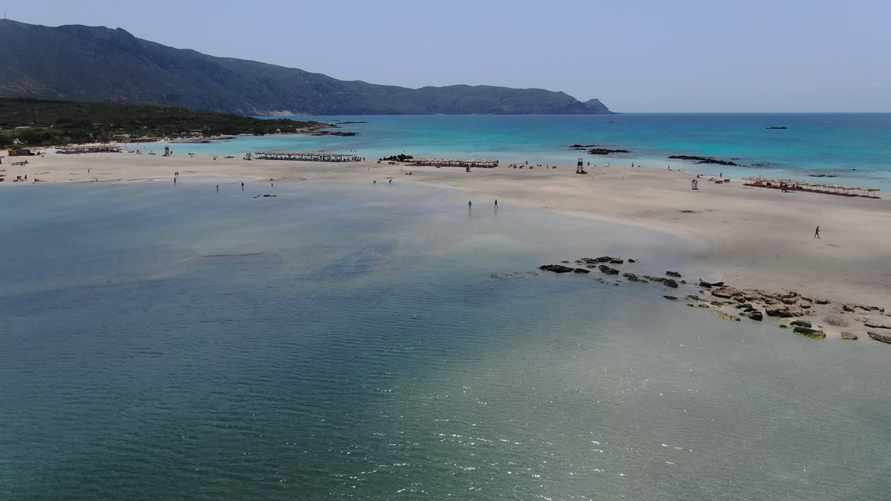 Elafonissi Beach saltwater lagoon in Crete Greece with bathers walking along the sand, Aerial dolly in shot