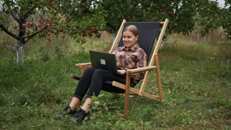 working remotely online via internet. young happy woman sits in the garden working on laptop and enjoying nature and fresh air. freelance job concept.