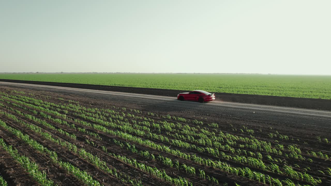Cinematic aerial following red colored sporting racing car moving along the road through vast agricultural fields, leaving dusty trail behind, daytime capture