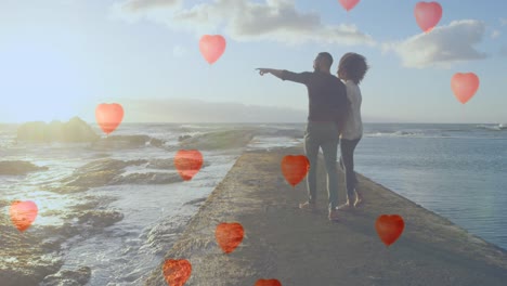 Multiple-heart-balloons-floating-against-couple-pointing-towards-sea-shore