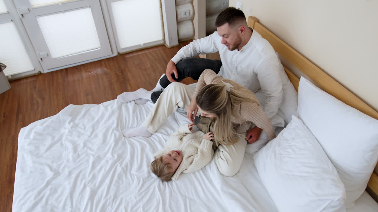 Caucasian couple sits on the big double bed hugging. Baby boy lying beside starts to cry and parents draw him closer. Mother calms down her son. Top view.