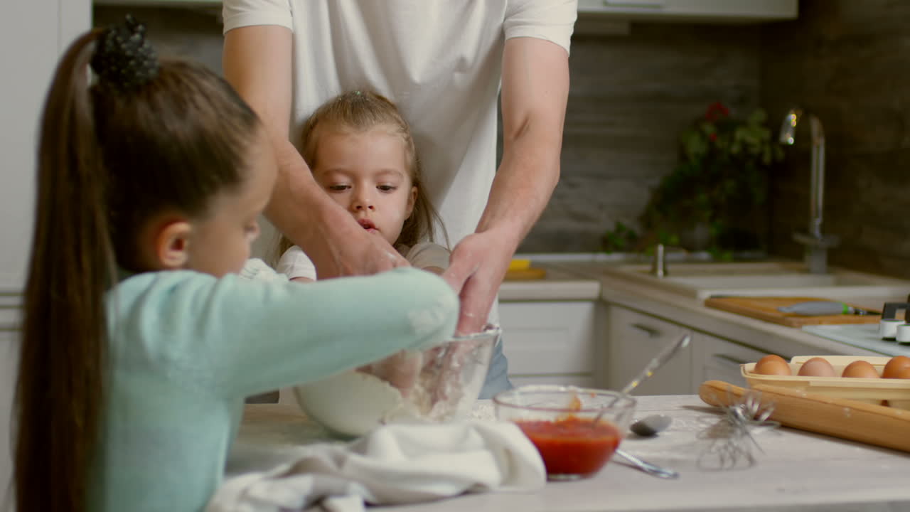 Zwei bezaubernde kleine Mädchen im Grundschulalter und ihr Vater kochen gemeinsam etwas und klatschen in die Hände, um das Mehl zu entfernen.