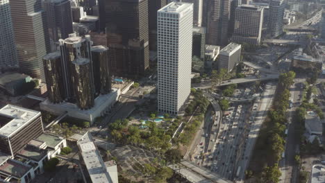 AERIAL:-Circling-Downtown-Los-Angeles,-California-intersection-traffic-with-palm-trees-and-Skyline-in-background-at-beautiful-blue-sky-and-sunny-day