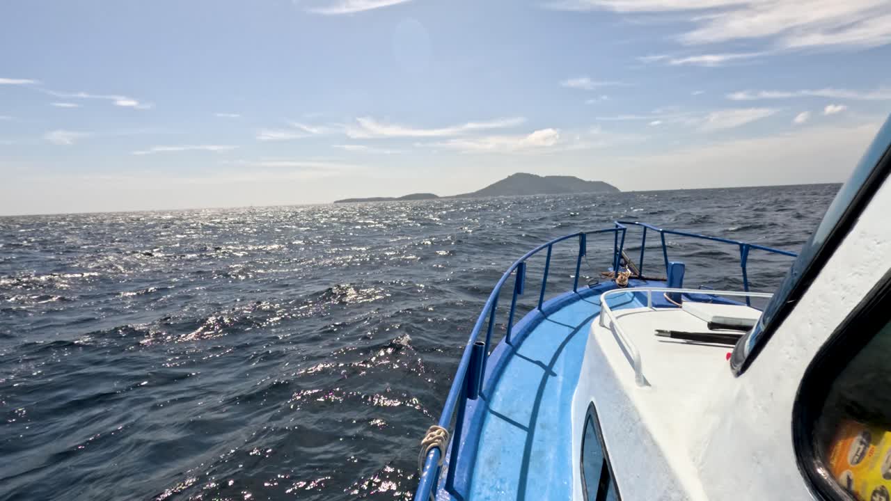 Blue boat moves across sparkling sea toward distant island, daylight, steady camera, Phuket, Thailand