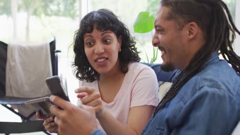 Happy-biracial-couple-in-living-room-using-laptop-and-talking,-with-her-wheelchair-in-background