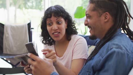 feliz pareja biracial en la sala de estar usando computadora portátil y hablando, con su silla de ruedas en el fondo