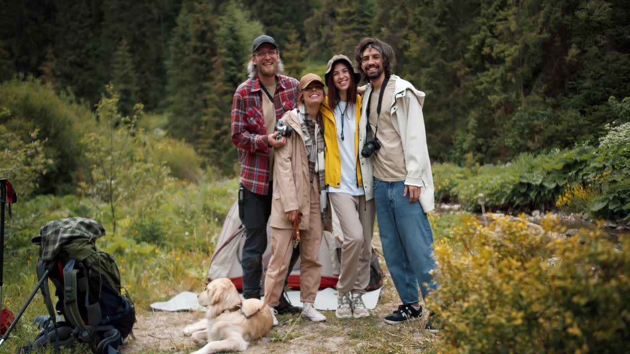 Happy company of tourists: 2 guys and 2 girls posing and looking at the camera in hiking clothes with backpacks against the backdrop of a green forest