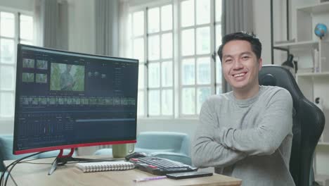 asian video editor man in long sleeved t-shirt and black pants crossing his arms and smiling due to successful editing video by desktop computer at home.