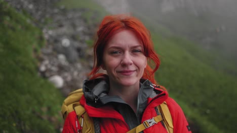 cámara lenta derecha panorámica de una niña excursionista sonriente, ella tiene el cabello rojo y lleva una chaqueta hinchada de color rojo naranja y una mochila amarilla