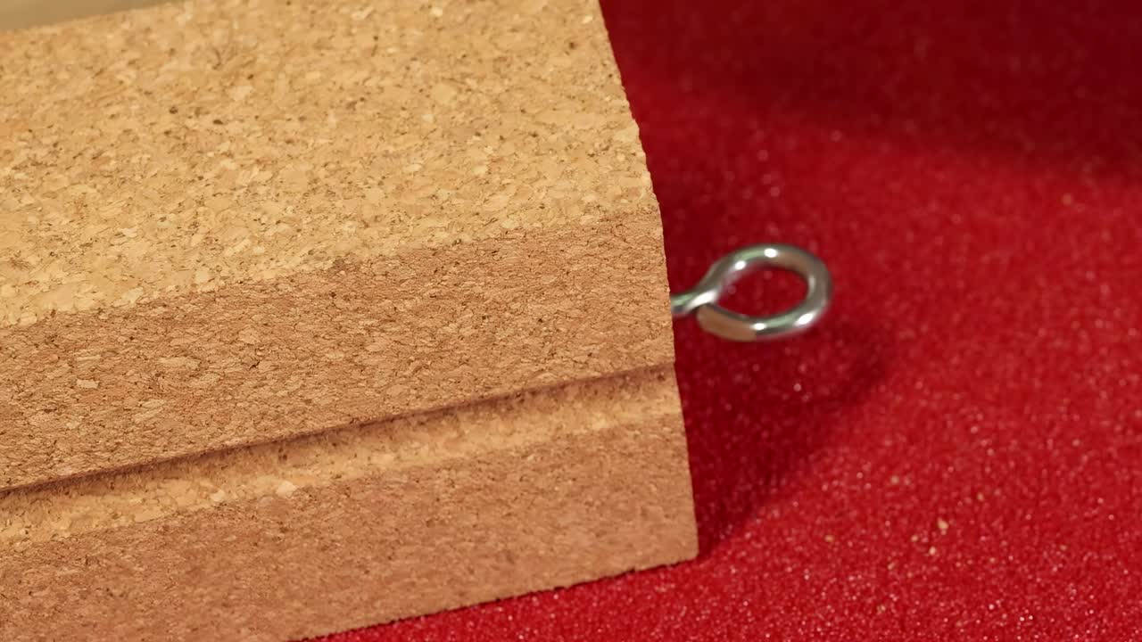A cork block with a metal hook is manipulated on a textured red surface using a blue tool.