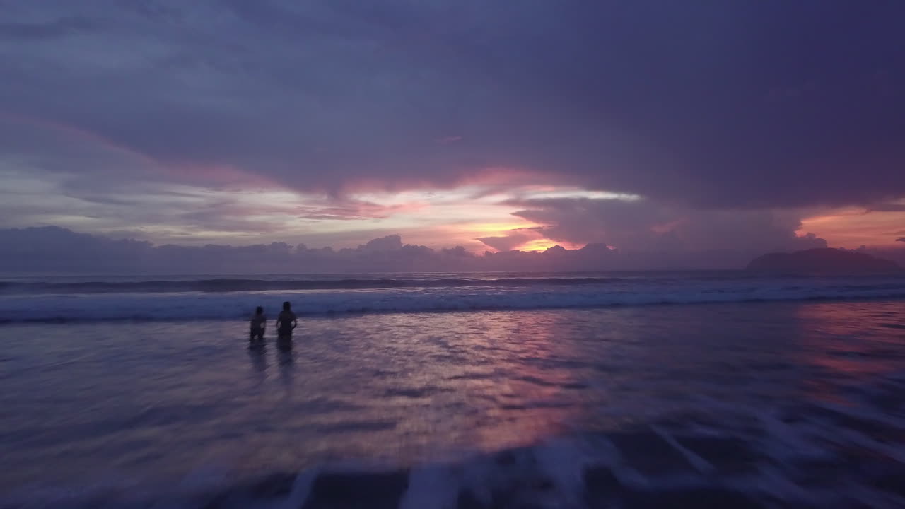 Dolly drone shot of a couple playing in the ocean on a beach in Jaco, Costa Rica during a beautiful sunset.