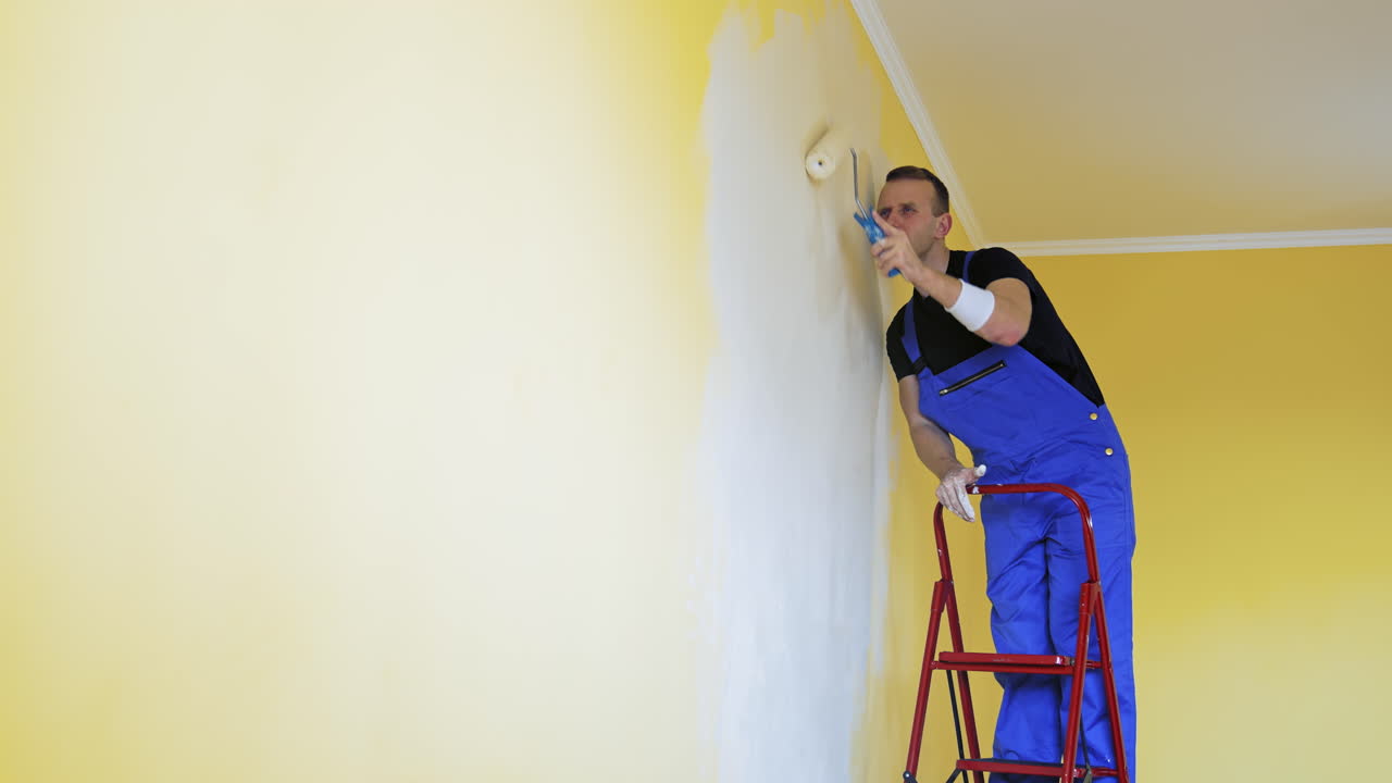 Young man in new apartment renovating room. Handsome painter working with brush roller renewing home.