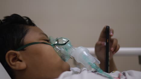 niño asiático con máscara de oxígeno en la cara en la cama del hospital mientras juega al teléfono celular