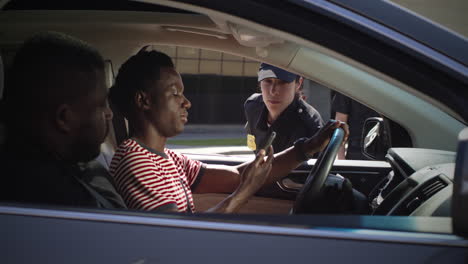 friendly police officer checking digital license of black driver zoom in view of positive woman in police uniform talking with african american man in car and scanning digital driver license on smartphone