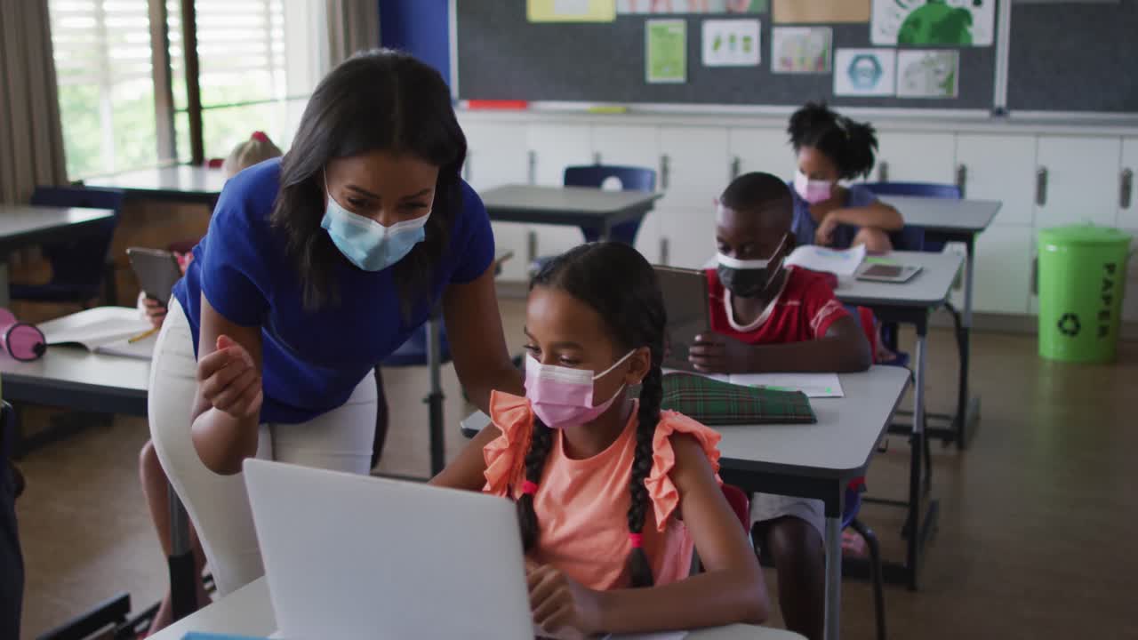 Insegnante donna multietnica che aiuta una scolara che usa il computer portatile, tutti con le mascherine.