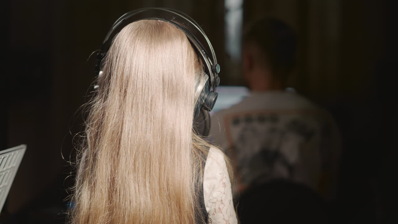 금발의 아이가 오디오 스튜디오에서 노래를 녹음합니다. 이어폰을 쓴 긴 머리 어린 소녀가 단열된 방에서 사운드 디렉터와 함께 마이크에서 노래합니다. 예술적 인 아이.