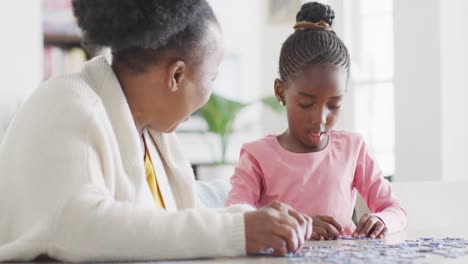 Happy-african-american-granddaughter-and-grandmother-doing-jigsaw-puzzle-at-home,-copy-space