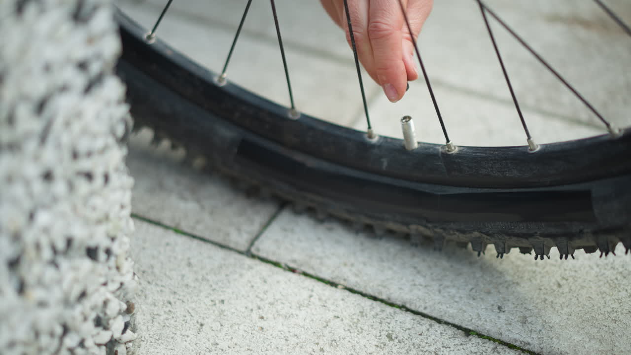 rimozione ravvicinata del cappuccio della valvola di una bicicletta da parte delle dita di una donna, marciapiede in pietra, cerchione sgonfio, polpastrello che solleva il cappuccio e il nucleo, movimento rapido e concentrato, pronto a gonfiare e continuare il tragitto, teso a sollevato