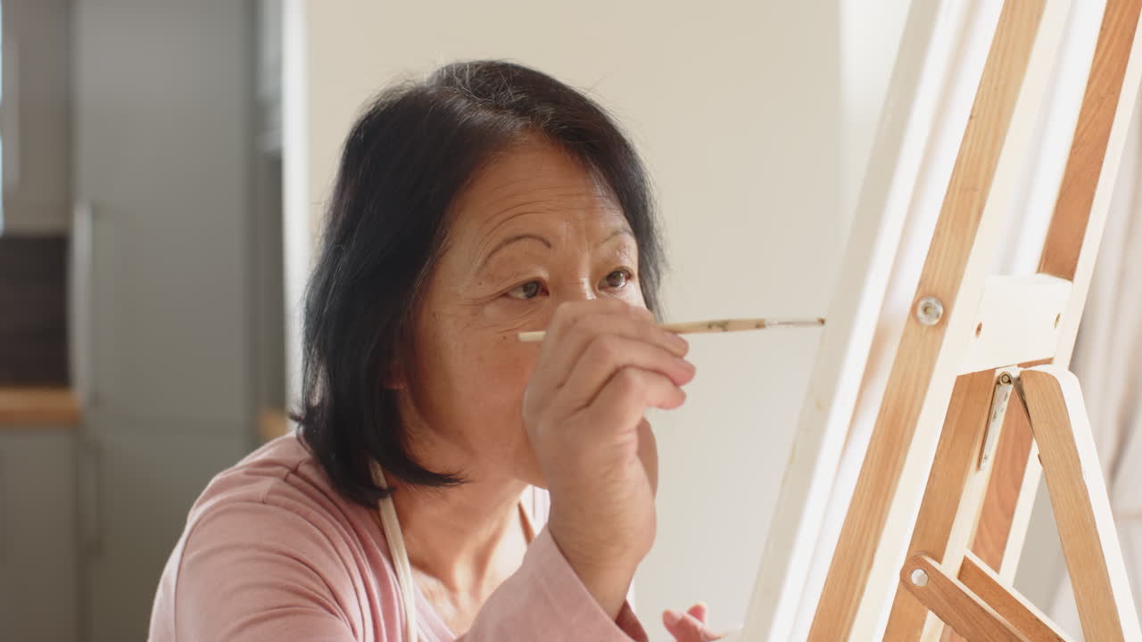 Senior Asian woman painting on canvas at home, focused and enjoying creative solitude