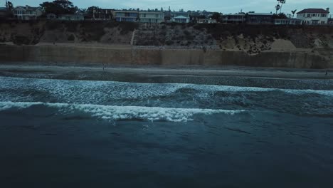 Blick-Von-Einer-Drohne,-Die-Nach-Links-über-Das-Meer-Fliegt-Und-Den-Strand-Und-Einige-Häuser-Am-Rand-Einer-Klippe-Zeigt